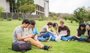 A group of teenagers with backpacks and touchscreen tablets sit in a half circle on a green lawn in front of a school building.